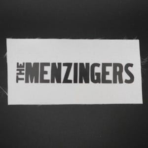 May include: A white rectangular patch with the text "THE MENZINGERS" in black, sans-serif font. The patch has a slightly frayed edge and is set against a black background. The text is centered on the patch.