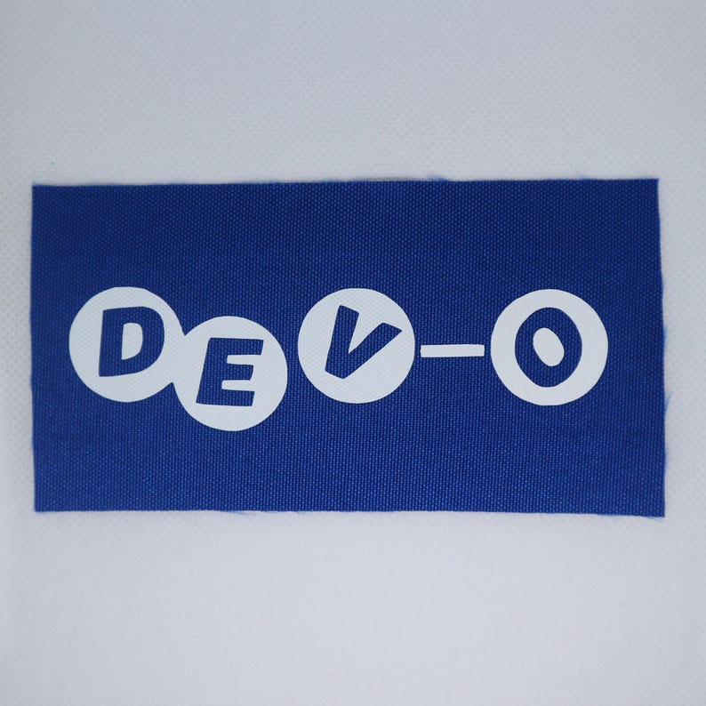 May include: A blue rectangular patch featuring the white text "DEVO-O". The letters are in a bold, rounded font. The patch has a textured appearance, and the background is a solid white.