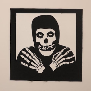 May include: A square black and white fabric patch featuring a high-contrast graphic illustration of a hooded figure with a skull face and skeletal hands crossed over the chest. The design is centered within a solid black border on a white background.