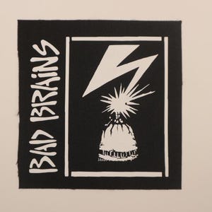 May include: Black square patch with white text and graphic. The text "BAD BRAINS" is vertically aligned on the left. A white lightning bolt and a stylized image are inside a white border.