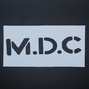 May include: A white rectangular stencil with the letters "M.D.C." in a bold, black, sans-serif font. The stencil is set against a black background. The letters are cut out.