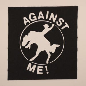 May include: Black square patch with white text and graphic. The text reads "AGAINST ME!" in a circular arrangement around a white silhouette of a person riding a bucking bronco. The patch has a rough, unfinished edge.