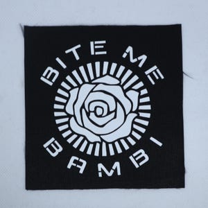 May include: A square black fabric patch featuring a white screen-printed design. The center displays a stylized rose surrounded by a sunburst pattern. The text BITE ME is arched above the rose, and the text BAMBI is arched below the rose.