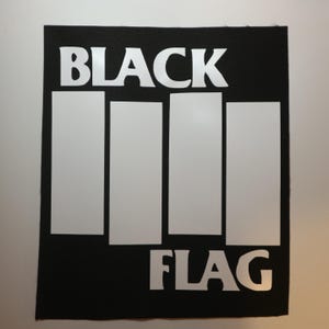 May include: A rectangular black fabric patch featuring the white text BLACK at the top and FLAG at the bottom. Between the words are four vertical white rectangular bars of varying heights, creating a high-contrast graphic design on a dark background.