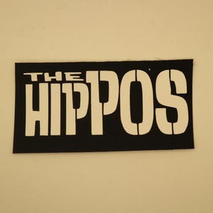 May include: A black rectangular patch with the white text "THE HIPPOS" in a stencil font. The patch is likely for clothing or accessories, featuring a bold, eye-catching design.