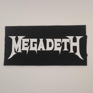 May include: Black rectangular patch with the white word "MEGADETH" in a stylized, gothic font. The patch has a textured surface and a simple, bold design, suitable for application on clothing or accessories.