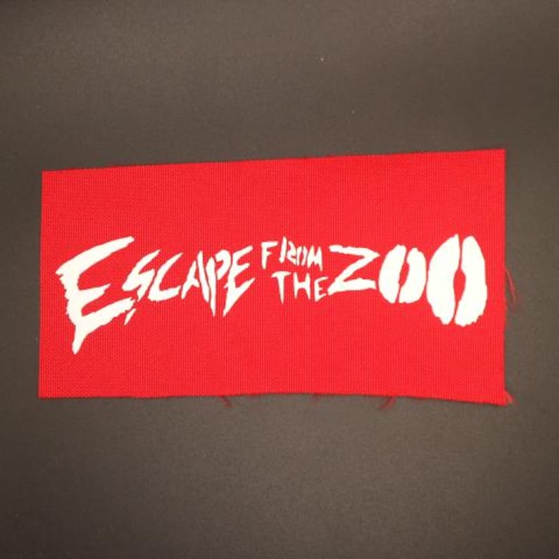 Op de afbeelding: Een rode rechthoekige patch met de witte tekst "ESCAPE FROM THE ZOO". De tekst heeft een verweerde, edgy lettertype. De patch is gemaakt van een geweven materiaal.