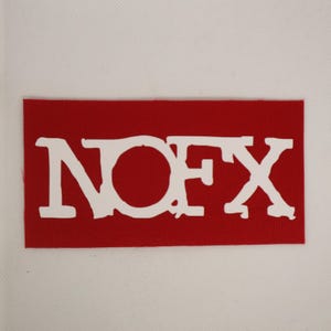 Peut inclure: Un &eacute;cusson rectangulaire rouge avec le texte blanc "NOFX" en gras, dans une police de caract&egrave;res vieillie. L'&eacute;cusson, probablement en tissu, est pos&eacute; sur un fond blanc. Les lettres sont l&eacute;g&egrave;rement irr&eacute;guli&egrave;res, donnant un aspect vintage.