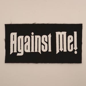 May include: Black rectangular patch with the words "Against Me!" in white, gothic-style lettering. The patch has a white background.