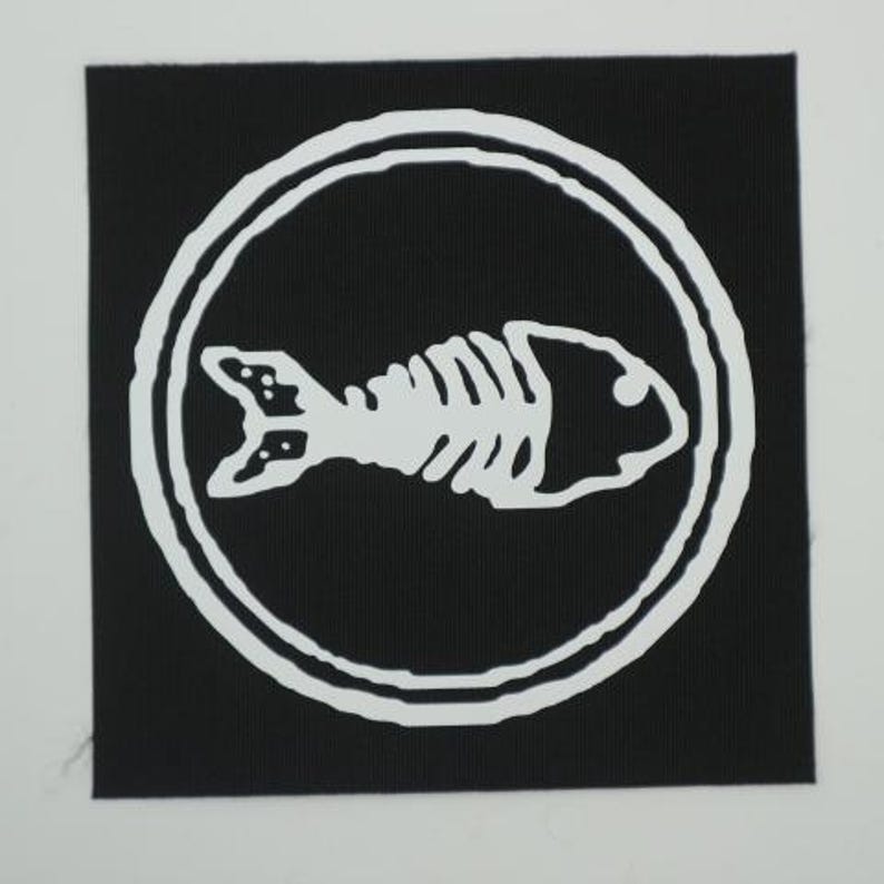 Puede incluir: Un parche cuadrado negro con un dise&ntilde;o de esqueleto de pez blanco dentro de un borde circular blanco. El esqueleto del pez est&aacute; estilizado, con costillas, cabeza y cola visibles. El parche es probablemente para ropa o accesorios.