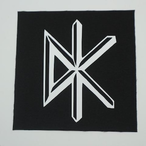 May include: A square black fabric patch featuring a white graphic logo in the center. The design consists of a stylized letter D combined with a letter K, rendered in a bold, angular, and geometric font with sharp, clean lines and edges.