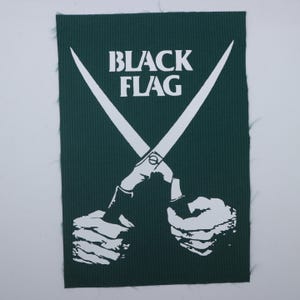 May include: A rectangular dark green fabric patch featuring a white graphic print of two hands holding a pair of crossed scissors. The text "BLACK FLAG" is printed in white capital letters above the scissors. The edges of the patch are frayed.