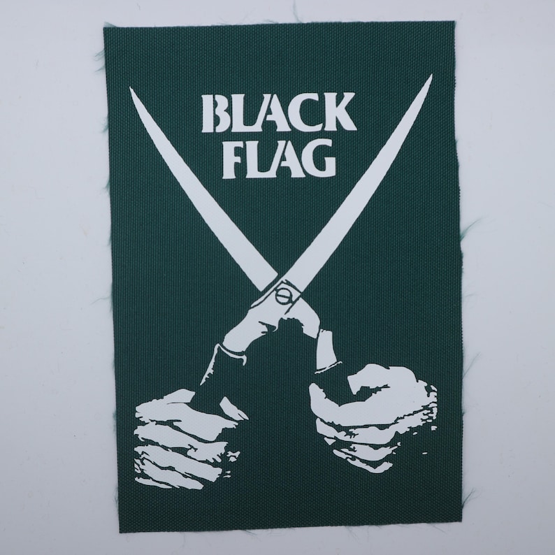 May include: A rectangular dark green fabric patch featuring a white graphic print of two hands holding a pair of crossed scissors. The text "BLACK FLAG" is printed in white capital letters above the scissors. The edges of the patch are frayed.