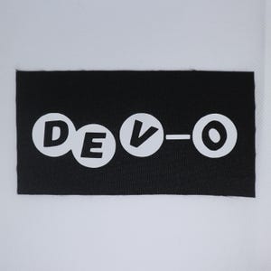 May include: A black rectangular patch featuring the word "DEVO" in white, circular lettering. The patch is designed for application on clothing or accessories.