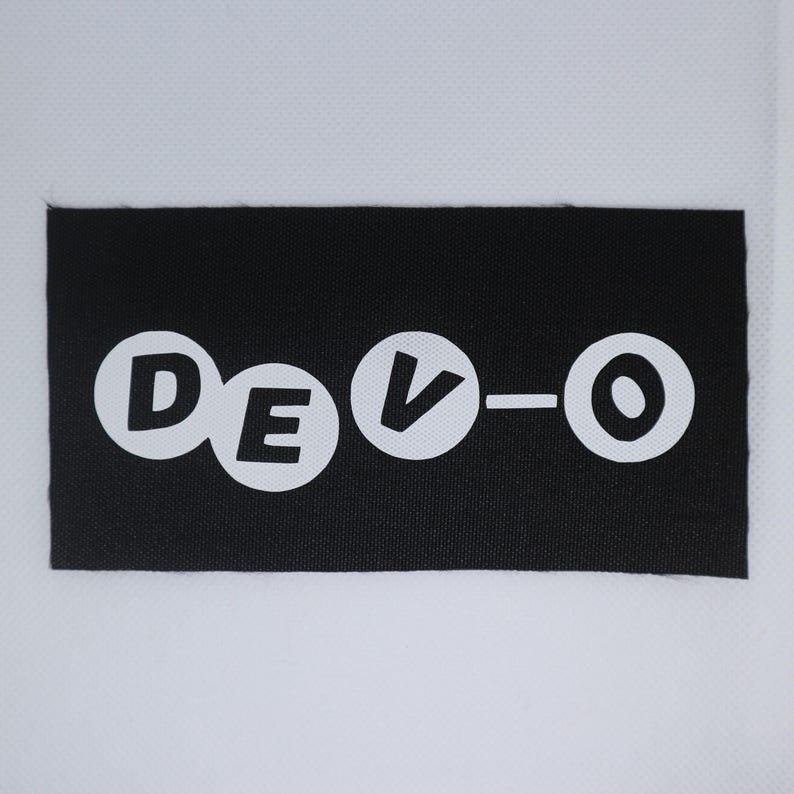 May include: A black rectangular patch featuring the word "DEVO" in white, circular lettering. The patch is designed for application on clothing or accessories.