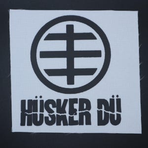May include: A white patch with a black circular emblem and the text "HÜSKER DÜ" in a bold, blocky font. The emblem features a cross-like shape within a circle, with three horizontal lines.