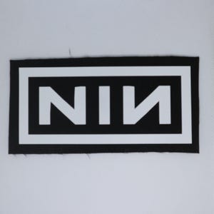 May include: A black rectangular patch with a white border and the letters "NIN" in white. The letters are in a bold, sans-serif font. The patch is likely for clothing or accessories.