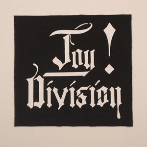 May include: A black square patch with the words "Joy Division" in white gothic lettering. The word "Joy" is above the word "Division". An exclamation point and a diamond shape are to the right of the word "Joy".