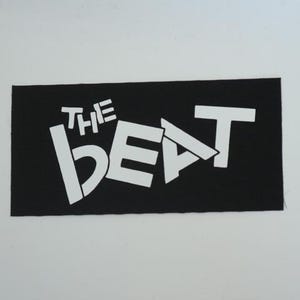 May include: A black rectangular patch with the white text "THE BEAT" in a bold, stylized font. The patch is likely made of fabric and is suitable for sewing or ironing onto clothing or accessories.