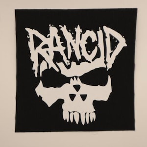May include: A square black fabric patch featuring a white screen-printed graphic. The design displays the word RANCID in a jagged, stylized font positioned above a skull graphic that incorporates a radiation symbol within the forehead area.