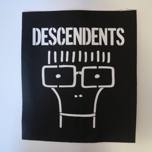 May include: Black square print featuring the word "DESCENDENTS" in white stencil font above a stylized face with glasses and hair. The graphic is a simple, bold design, perfect for fans of the band.