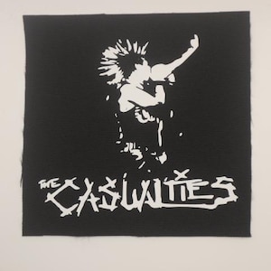 May include: Black square patch featuring a white graphic of a person with a mohawk and raised arm, with the band name "THE CASUALTIES" in white lettering below. The patch has a raw edge.