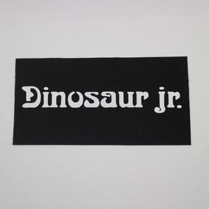 May include: A black rectangular patch with the white text "Dinosaur jr." in a stylized font. The patch has a textured surface and is set against a white background. The text is the primary focus of the image.