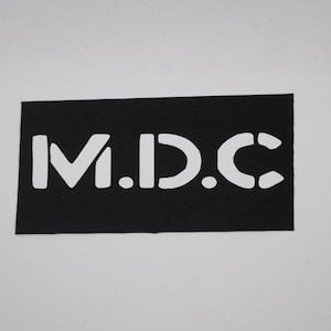 May include: A black rectangular patch with the white letters "M.D.C." in a stencil font. The patch is likely made of fabric and is set against a plain white background.
