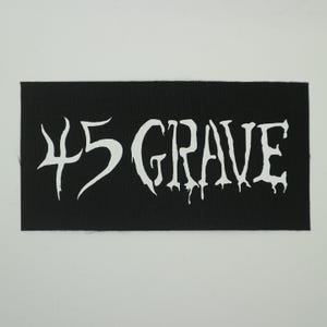 May include: Black rectangular patch with the white text "45 GRAVE" in a stylized, dripping font. The patch has a woven texture and is set against a plain white background. The text is the focal point.