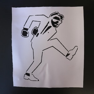 May include: Black and white stencil art of a man in a suit, hat, and sunglasses, in a walking pose. The image is on a white fabric, with a black background. The man is wearing a suit, tie, and shoes.
