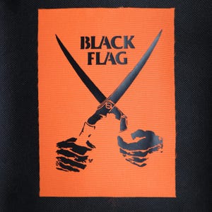 May include: A rectangular fabric patch with a bright orange background and a black graphic print. The design features two crossed blades held by two hands, with the text BLACK FLAG printed in bold black capital letters above the blades.