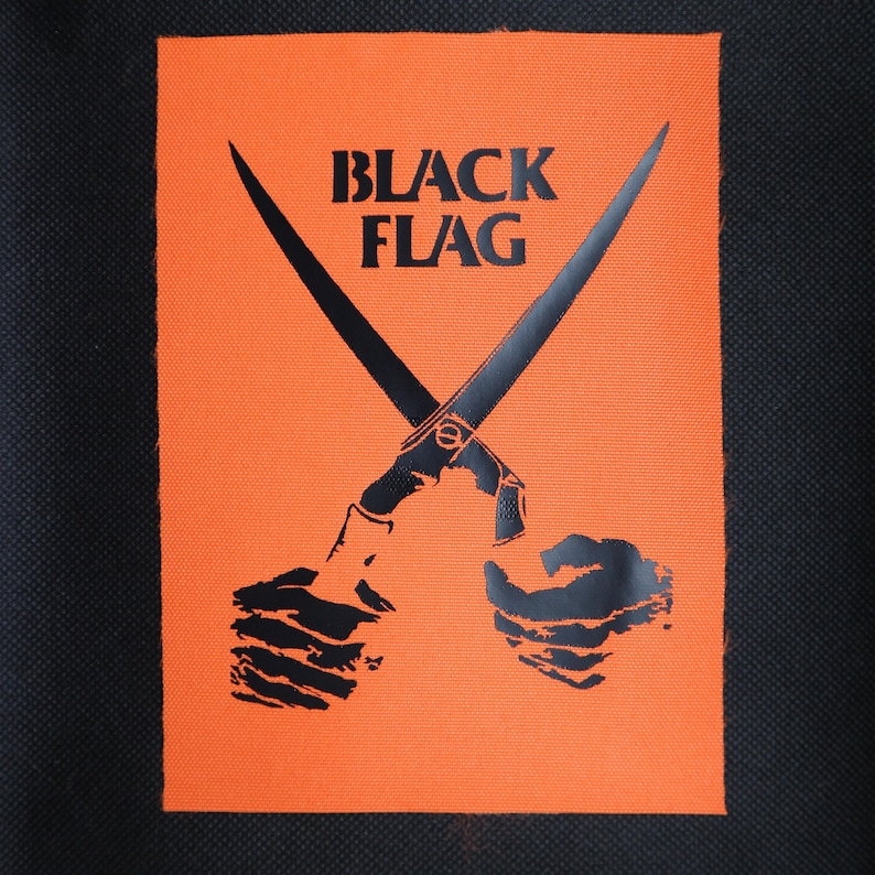 May include: A rectangular fabric patch with a bright orange background and a black graphic print. The design features two crossed blades held by two hands, with the text BLACK FLAG printed in bold black capital letters above the blades.