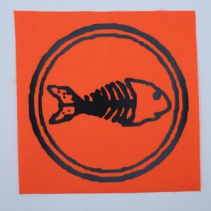 Puede incluir: Parche cuadrado naranja con un dise&ntilde;o de esqueleto de pez negro dentro de un borde circular negro. El esqueleto del pez es una ilustraci&oacute;n estilizada. El parche tiene una superficie texturizada.