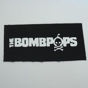 May include: A rectangular black fabric patch featuring the white text THE BOMBPOPS. The letter O in BOMBPOPS is replaced by a white graphic of a skull with a lit fuse on top, positioned above a pair of crossed bones, creating a skull and crossbones icon.
