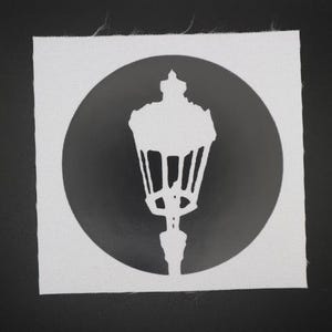 May include: A white square fabric patch featuring a black silhouette of a vintage street lamp inside a black circle. The lamp has a detailed design with a decorative top and a slender base. The background is a solid black.