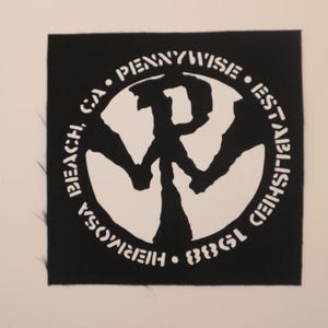 May include: A black square patch with a white circular design and text. The design features a stylized letter "P" within a circle, with the words "PENNYWISE ESTABLISHED" and "HERMOSA BEACH, CA 1988" encircling it. The patch has a raw edge.
