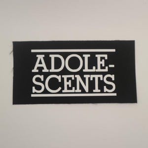 May include: Black rectangular patch with the word "ADOLESCENTS" in bold white capital letters. The text is centered and framed by horizontal white lines. The patch has a slightly frayed edge.