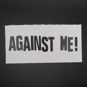 May include: A rectangular patch with the words "AGAINST ME!" printed in bold, black letters. The text is centered on a white, textured fabric, set against a dark background.