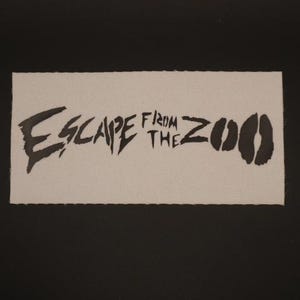 Op de afbeelding: Een rechthoekige patch met de woorden "ESCAPE FROM THE ZOO" in zwarte, gestileerde letters. De patch heeft een lichtbeige achtergrond en een zwarte rand. Het ontwerp bevat twee zwarte dierenvoetsporen.