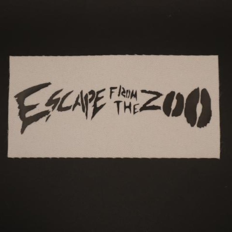 Op de afbeelding: Een rechthoekige patch met de woorden "ESCAPE FROM THE ZOO" in zwarte, gestileerde letters. De patch heeft een lichtbeige achtergrond en een zwarte rand. Het ontwerp bevat twee zwarte dierenvoetsporen.