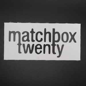 May include: A rectangular white fabric patch featuring the text matchbox twenty printed in a dark gray, sans-serif font. The patch has a visible woven texture and clean, straight edges, presenting the band name clearly across two lines.