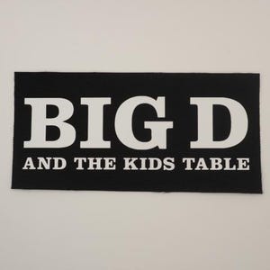 May include: A black rectangular patch with white text. The text reads "BIG D" in large capital letters, with "AND THE KIDS TABLE" in smaller capital letters below. The patch is likely for clothing or accessories.
