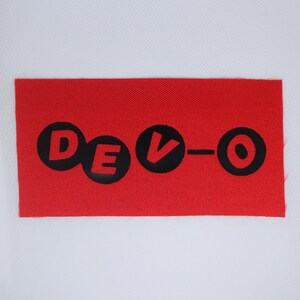 May include: A red rectangular patch featuring the word "DEVO" in black, rounded letters. The "V" has a downward-pointing arrow. The patch is made of a textured fabric. The design is simple and bold.