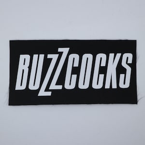May include: A black rectangular patch with the white text "BUZZCOCKS" in a bold, sans-serif font. The patch has a woven texture and is likely designed to be sewn onto clothing or accessories.