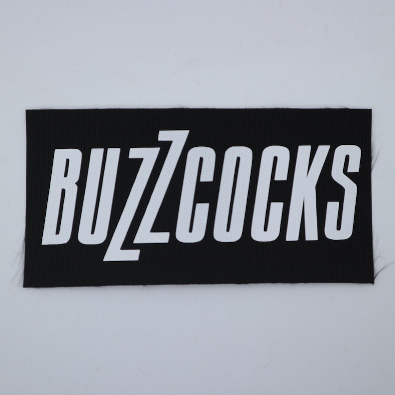 Op de afbeelding: Een zwart rechthoekig embleem met de witte tekst "BUZZCOCKS" in een vetgedrukte, schreefloze letter. De patch heeft een geweven textuur en is waarschijnlijk bedoeld om op kleding of accessoires te worden genaaid.