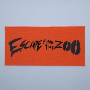 Op de afbeelding: Een oranje rechthoekige patch met de zwarte woorden "ESCAPE FROM THE ZOO" in gestileerde letters. Het woord "ZOO" wordt gevolgd door twee zwarte dierenpootafdrukken.