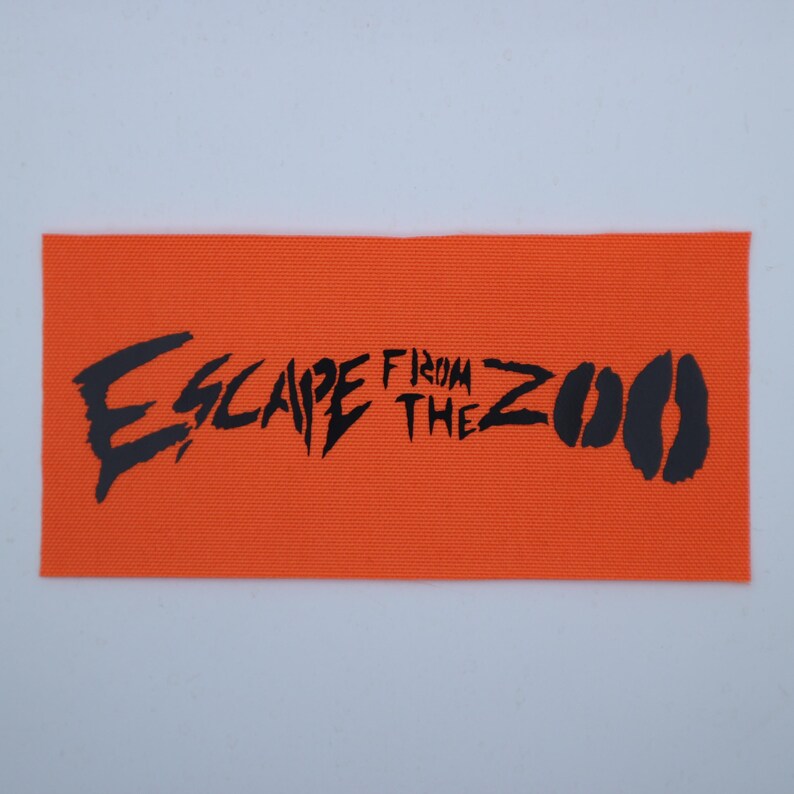Op de afbeelding: Een oranje rechthoekige patch met de zwarte woorden "ESCAPE FROM THE ZOO" in gestileerde letters. Het woord "ZOO" wordt gevolgd door twee zwarte dierenpootafdrukken.