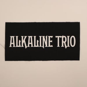 May include: A black rectangular patch with the white text "ALKALINE TRIO" in a stylized font. The patch is likely made of fabric and is set against a plain, off-white background. The text is the focal point.