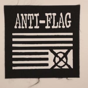 May include: Black square patch with the words "ANTI-FLAG" in white block letters above a white graphic resembling a flag with a circle and cross through it. The patch has a rough, frayed edge.