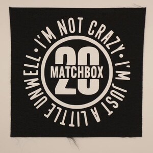 May include: A square black fabric patch featuring a white circular graphic. The design includes the large number 20, the word MATCHBOX, and the text I'M NOT CRAZY I'M JUST A LITTLE UNWELL arranged in a circle around the center elements.